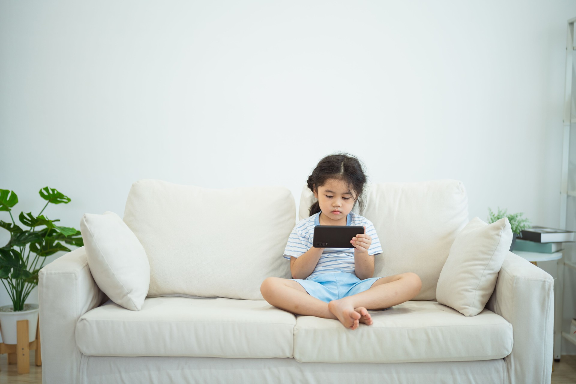 Asian child girl looking using and touch mobile phone screen on couch sofa. Baby smiling funny time to use mobile phone. Too much screen time. Cute girl watching videos while tv, Internet addiction.