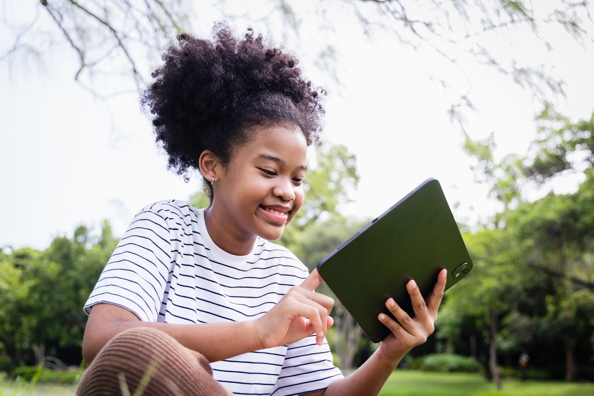 Happy African American girl sitting on grass by the lake, holding a digital tablet with stylus, focusing on drawing and learning in a calm and natural park environment