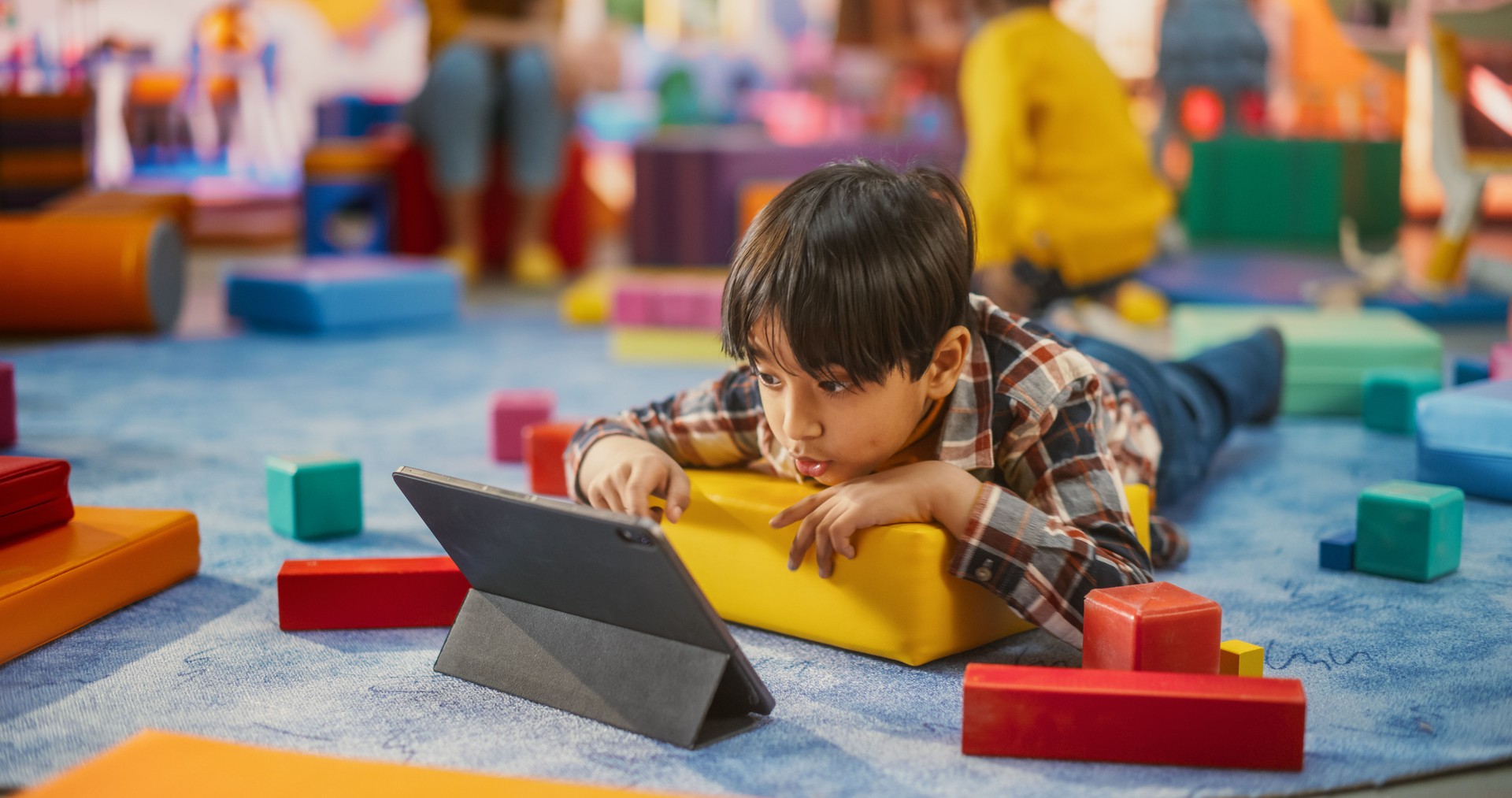 Talented Asian Boy Lying on the Floor and Using a Tablet Computer for Studying and Playing in Kindergarten. Handsome Young Child Spending Time with Technology in a Modern Daycare Center